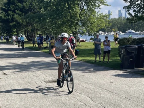 Male young adult riding his bicycle with focus and determination down a paved street with spectators watching from the edge of the road.