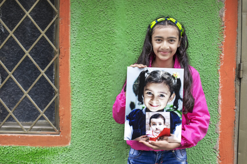 Young girl holds two photos of herself at younger ages in front of a green wall.