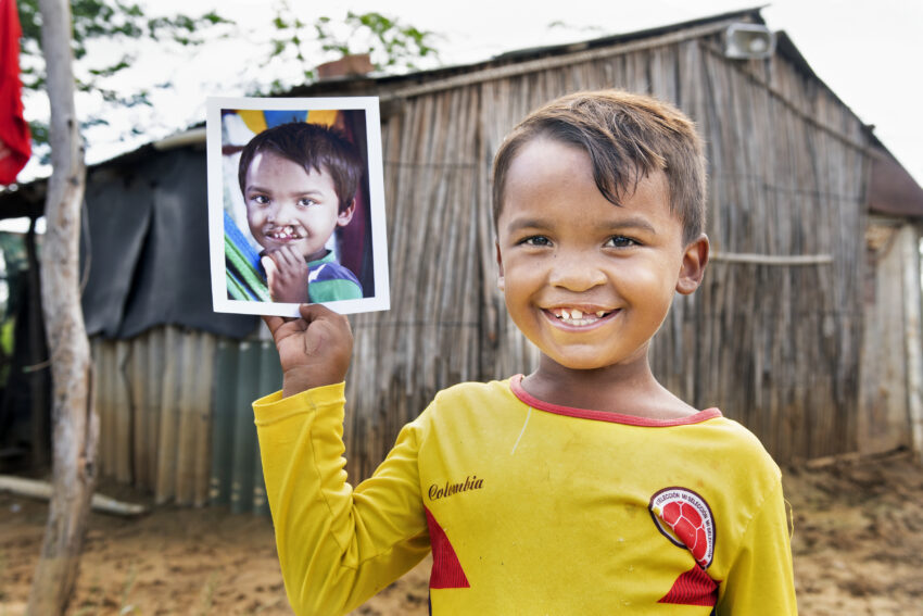 Young boy is holding a photo of himself in front of his wooden home.