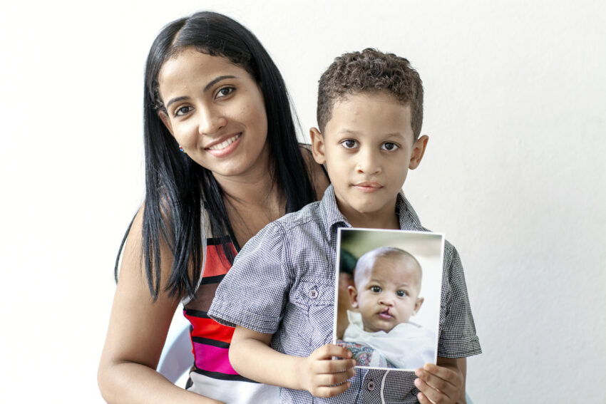 Mother smiles next to her child who is a young boy holding a photo of his younger self before receiving cleft surgery.