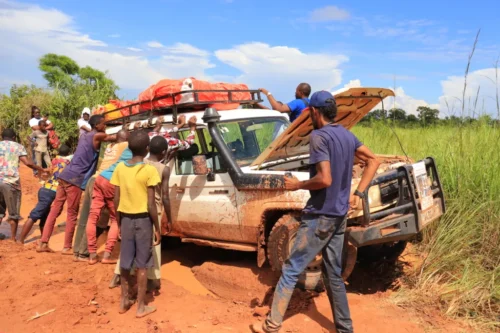 A vehicle with its hood up is parked in thick, orange mud. Several people, of all ages, stand around it with hands leaning on it.
