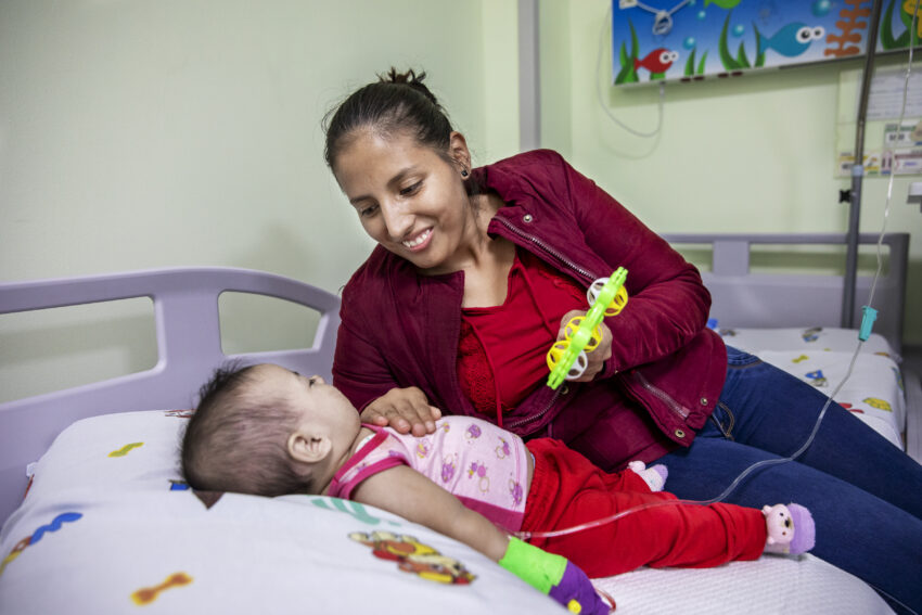 Mother holds a toy as she comforts her baby who is lying in a hospital bed.