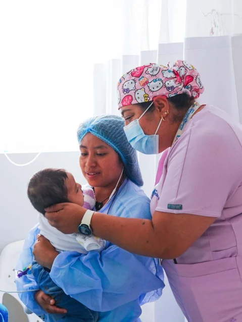 A nurse caringly holds a young patient's head while the mother watches her child's face after surgery.