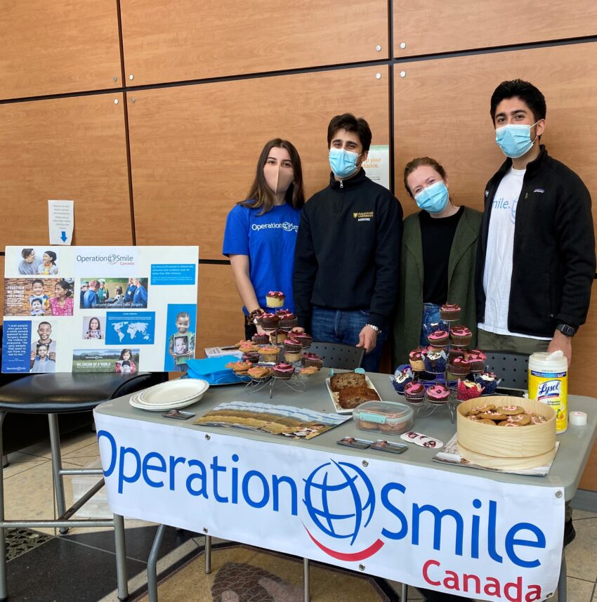 Four students in face masks running a bake sale for Operation Smile Canada.