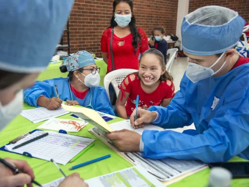 A teenage girl smiles brightly while surrounded by three medical volunteers who are determining whether she will receive cleft lip surgery. Her mother stands behind her, smiling behind her face mask, knowing her daughter's life is about to change.