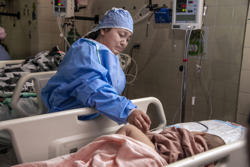 mother dressed in surgical scrubs looks at her son in a hospital bed with happy tears in her eyes