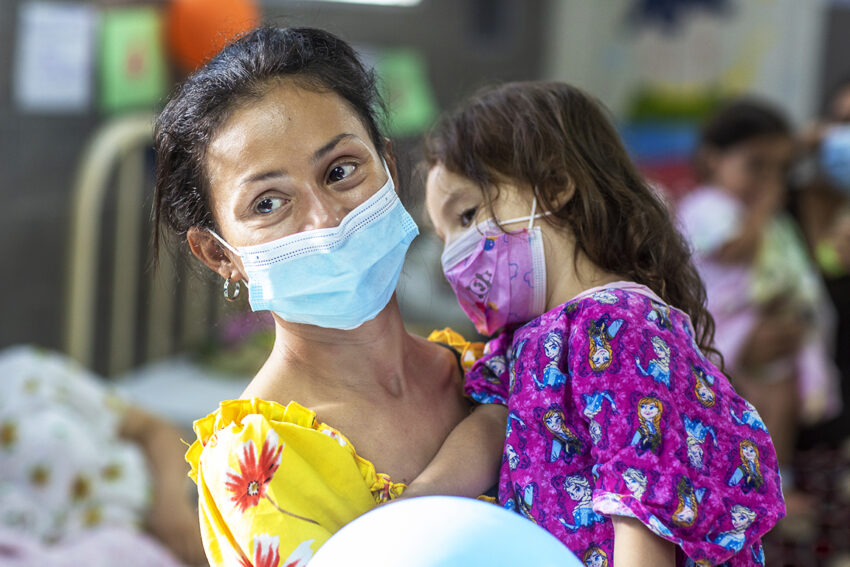 A mother holds her toddler daughter who is dressed in a hospital gown.