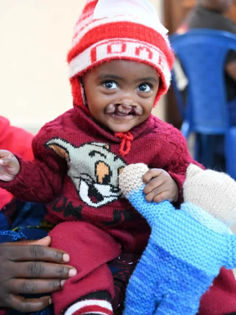 A young child wearing knit clothes is smiling brightly after being handed a teddy bear