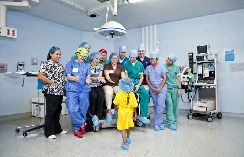 A room full of medical professionals smile and clap as a child in a hospital gown walks towards them in an OR. 