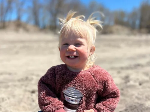 Toddler girl with white blonde hair tied in pig tails sitting in the sand, smiling proudly at the camera.